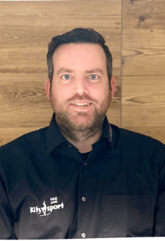 Porträt eines Mannes mit Bart, der ein Kitz Sport-Shirt vor einem Holzpaneel-Hintergrund trägt.