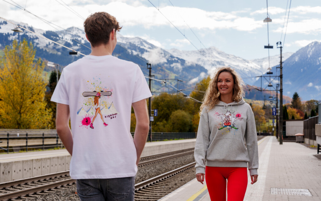 Mann in weißem Grafik-T-Shirt, Frau in grauem Grafik-Hoodie auf dem Bahnsteig, verschneite Bergkulisse.