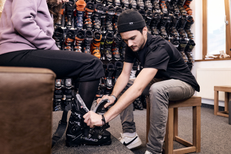 Skiverkäufer passt einem Kunden Skischuhe an, mit Reihen von Skischuhen im Hintergrund.