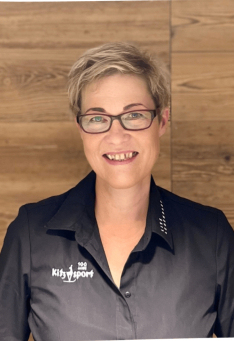 Lächelnde Frau mit Brille, die ein Kitz Sport Shirt vor einer holzgetäfelten Wand trägt.