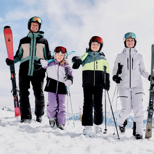 Eine vierköpfige Familie in bunter Skiausrüstung steht auf einem verschneiten Berg, bereit für den Wintersport.
