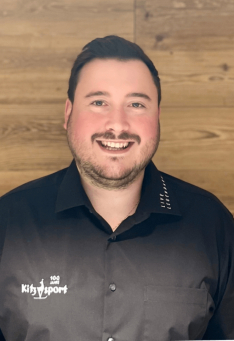 Porträt eines Kitzsport-Mitarbeiters in einem schwarzen Uniformhemd vor einem Holzpaneel-Hintergrund.