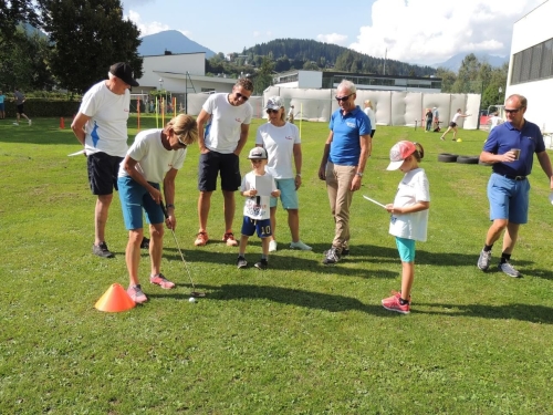 Eine Gruppe von Leuten spielt Golf auf einem sonnigen, grünen Platz mit Bergkulisse.