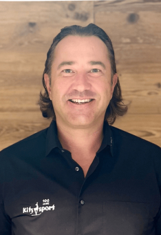 Porträt eines Kitzsport-Mitarbeiters im schwarzen Shirt vor Holzpaneel-Hintergrund.