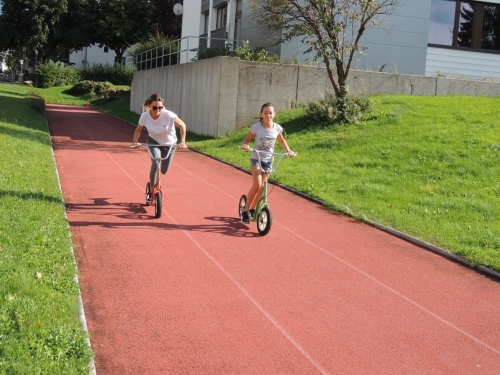 Zwei Mädchen fahren Roller auf einer roten Bahn im Freien in einer Parkanlage.