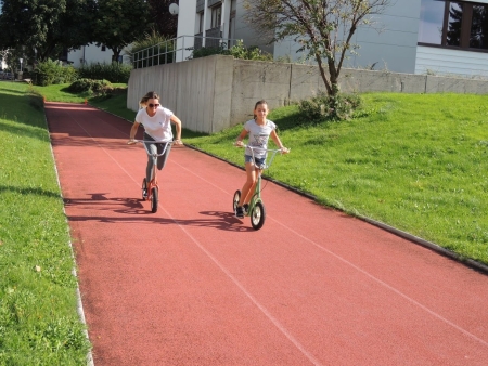 Zwei Mädchen fahren Roller auf einer roten Bahn im Freien in einer Parkanlage.