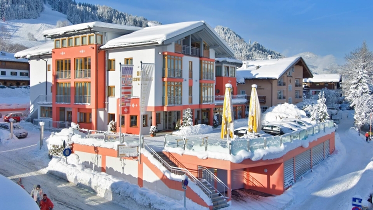 Schneebedecktes österreichisches Alpenresorthotel mit orangefarbenen Akzenten, Sonnenschirmen und Bergblick.