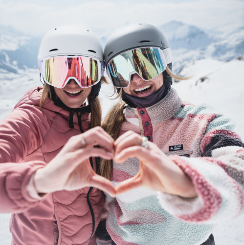 Zwei lächelnde Frauen in Skihelmen und verspiegelten Skibrillen bilden auf einem verschneiten Berg eine Herzform für einen Winterskiurlaub.