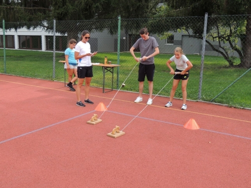 Kinder nehmen an einem Holzski-Rennen auf einer roten Piste unter Aufsicht eines Skilehrers teil.