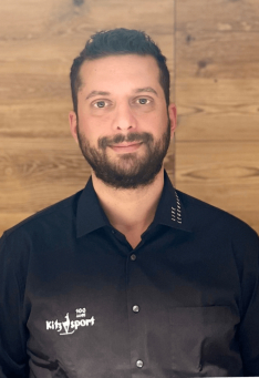 Porträt eines Kitz Sport Mitarbeiters im schwarzen Hemd vor einer Holzvertäfelung.