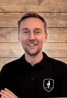 Porträt eines lächelnden Mannes in schwarzem Poloshirt vor einem Holzpaneel-Hintergrund.