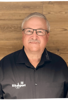 Porträt eines Mannes mit Brille und Kitzsport-Shirt vor einem Hintergrund mit Holzvertäfelung.