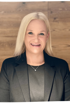 Professionelles Porträt einer blonden Frau in einem schwarzen Blazer vor einem Holzpaneel-Hintergrund.