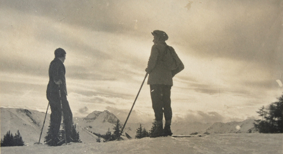 Vintage-Foto: Zwei Skifahrer mit Stöcken auf schneebedecktem Berggipfel mit Blick auf die Alpen.