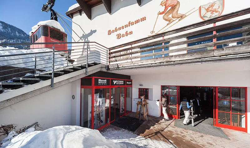 Skigebiets-Eingang mit roter Gondelbahn, Schnee und einem Wandbild mit Skifahrern in Serfaus-Fiss-Ladis, Österreich.