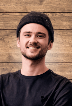 Lächelnder junger Mann mit Mütze und schwarzem T-Shirt vor einem rustikalen Holzpaneel-Hintergrund. Porträt.