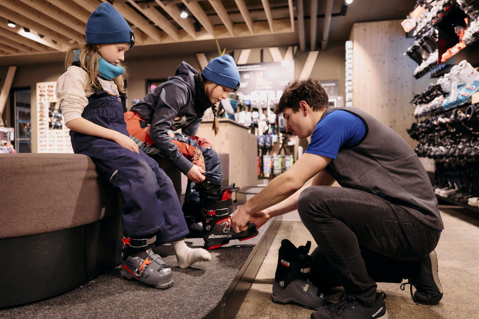 Ein Mitarbeiter eines Skigeschäfts passt zwei kleinen Kindern Skischuhe für die Wintersportausrüstung an.